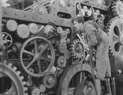 Fotograma de la película [Tiempos modernos](https://es.wikipedia.org/wiki/Tiempos_modernos) (1936), donde aparece [Charles Chaplin](https://es.wikipedia.org/wiki/Charles_Chaplin), vestido de trabajador, subido a una escalera mirando con asombro al mecánico que parece surgir de la propia máquina repleta de engranajes.
Fuente: https://ezer1film.blog.hu/2017/04/06/95_modern_idok_modern_times.